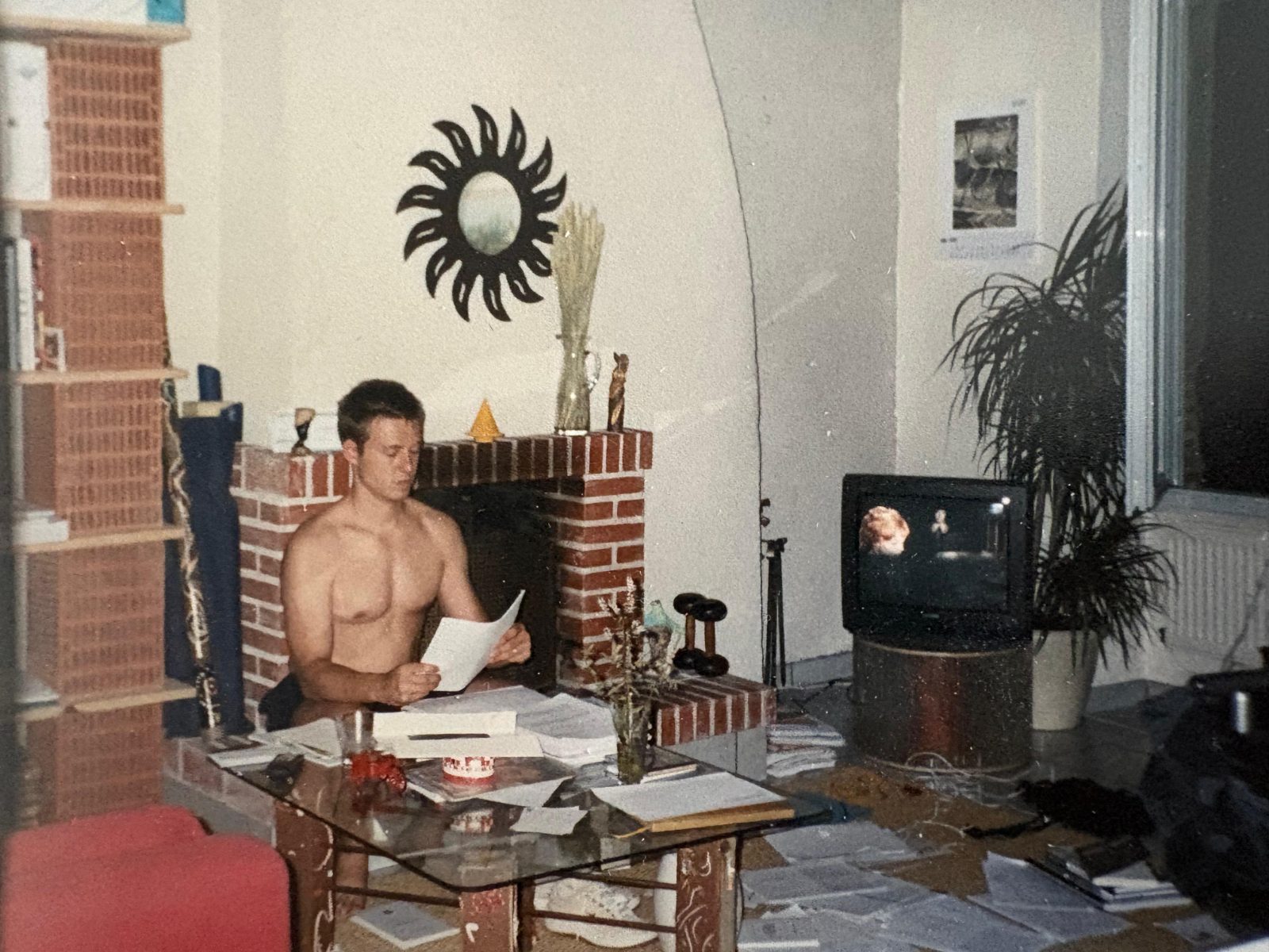 A print from the thesis years: Nicolas Boitout reading a sheet of
                          paper at a glass table in front of a brick fireplace, with books,
                          notes and loose pages spread over the table and across the floor
                          around him.